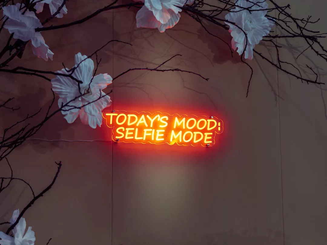 Ein Neonschild mit der Aufschrift "TODAY'S MOOD: SELFIE MODE" an einer Wand, umgeben von Zweigen mit weißen Kunstblumen - eine auffällige, von GLAMESiA inspirierte Kulisse.