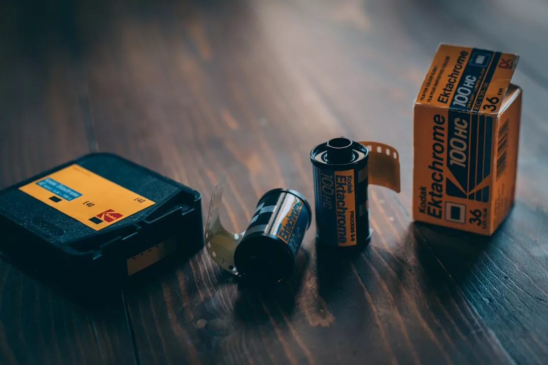 Drei Kodak Ektachrome 100HC-Filmrollen, eine in einer schwarzen Hülle, und eine gelbe Filmdose werden auf einer hölzernen Oberfläche präsentiert und fangen die zeitlose Atmosphäre der analogen Fotografie mit einem Hauch von GLAMESiA-Flair ein.