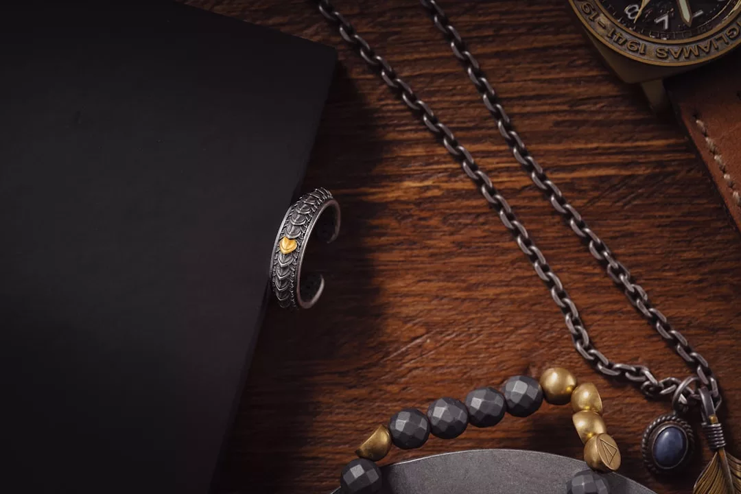 Ein silberner Ring mit goldenen Akzenten von GLAMESiA ruht auf einer schwarzen Fläche neben einer Metallkette, einem Perlenarmband und einem Teil einer Armbanduhr auf einem Holztisch.