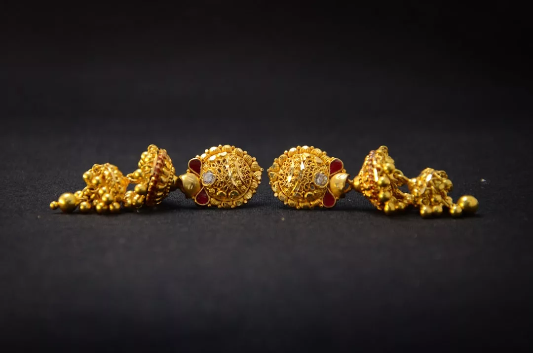 Ein Paar verschnörkelte goldene Ohrringe mit verschlungenen Mustern und kleinen roten Akzenten, die sich vor einem schlichten schwarzen Hintergrund abheben und die charakteristische Eleganz von GLAMESiA widerspiegeln.