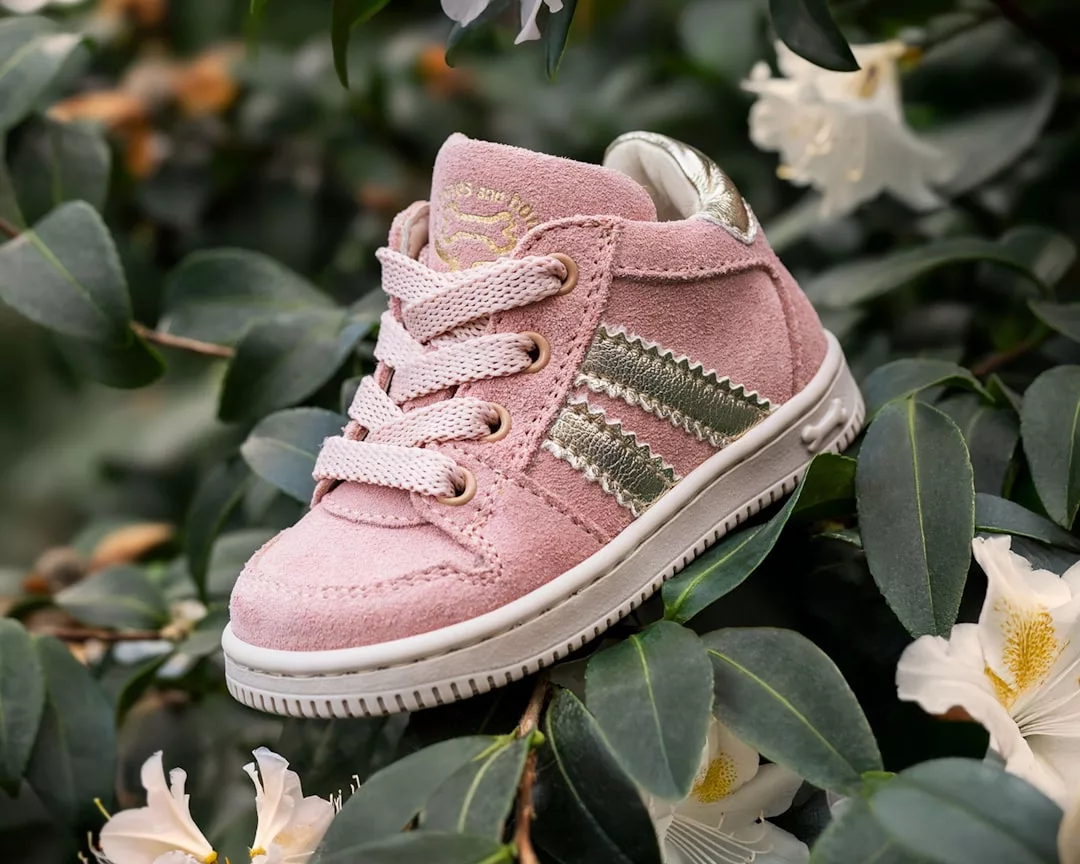 Ein rosa-goldener GLAMESiA-Kleinkind-Sneaker mit weißen Schnürsenkeln und Gummisohle wird auf grünem Laub mit weißen Blumen präsentiert.