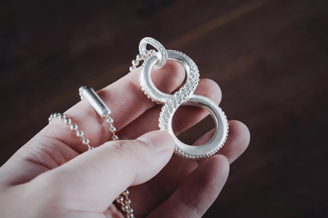 Eine Hand hält eine silberne GLAMESiA-Halskette mit einem Anhänger in Form eines Unendlichkeitssymbols, das mit einem strukturierten Saugnapfmuster versehen ist, das an einen Krakententakel erinnert.