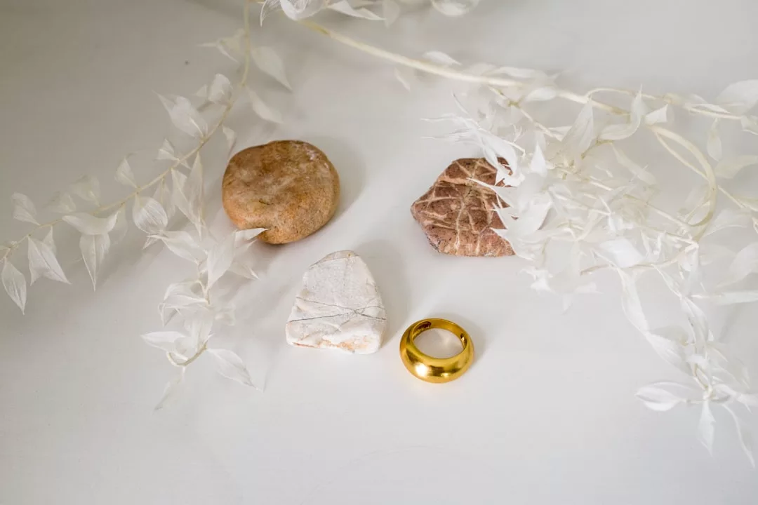 Drei Steine und ein Goldring von GLAMESiA sind auf einer weißen Fläche angeordnet, wobei blasse, getrocknete Blätter die Objekte teilweise von oben einrahmen.