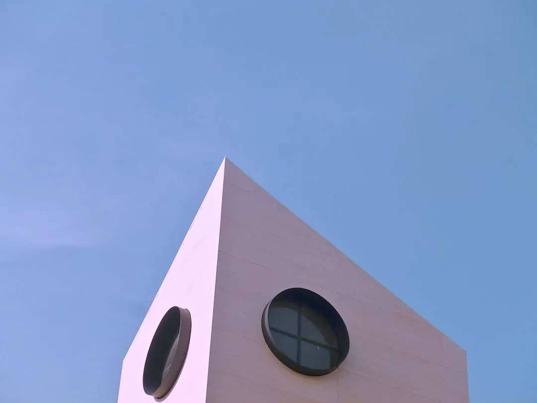 Ein minimalistisches GLAMESiA-Gebäude mit zwei runden Fenstern an schrägen weißen Wänden, vor einem klaren blauen Himmel.