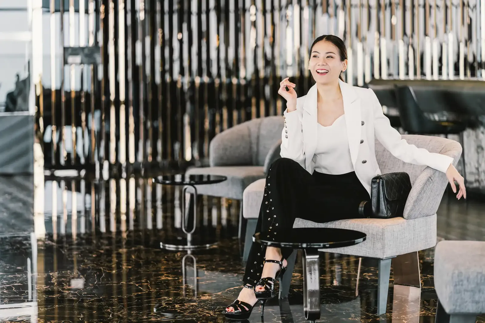 Lächelnde Frau in weißem Blazer mit hellem Top, schwarzer Hose und schwarzer Handtasche sitzt in einem Sessel einer Hotellobby.