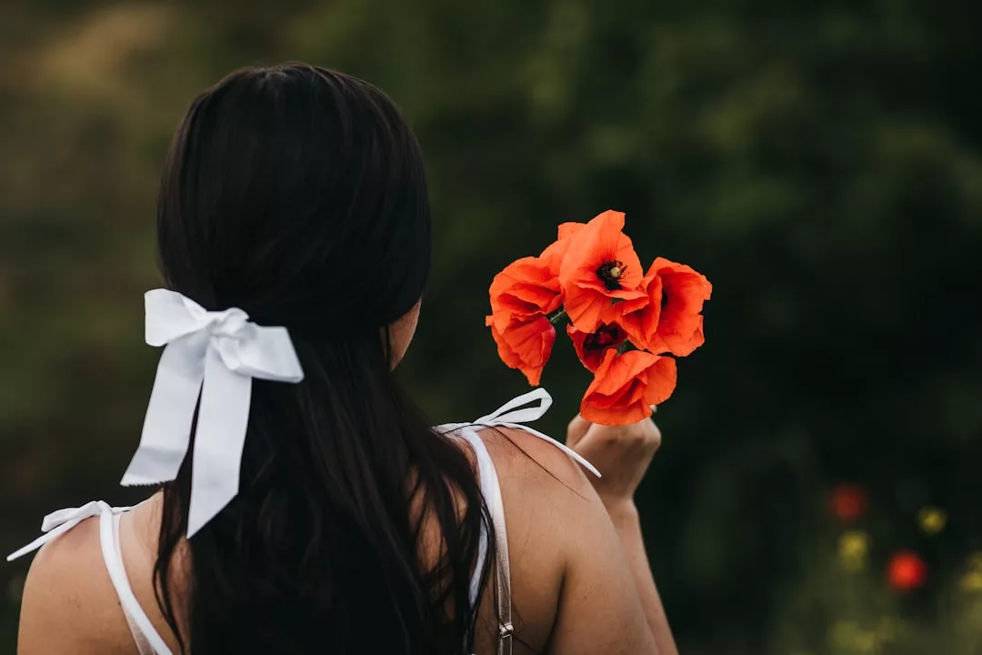 Eine Frau mit langen dunklen Haaren, die mit einem weißen Band zusammengebunden sind, hält zwei leuchtend orangefarbene Blumen in der Hand. Sie steht im Freien mit Grünpflanzen im Hintergrund und verkörpert die Eleganz und den Charme von GLAMESiA.