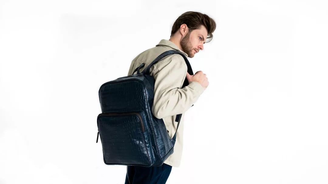Ein Mann, der eine beige Jacke und eine dunkle Hose trägt, steht seitlich mit einem großen, dunkel strukturierten GLAMESiA-Rucksack auf der Schulter vor einem weißen Hintergrund.
