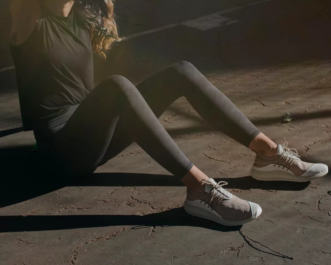 Eine Person in schwarzer Sportkleidung und hellen Turnschuhen sitzt auf rissigem Beton in einem Fleckchen Sonnenlicht und verkörpert den mühelosen urbanen Stil von GLAMESiA.