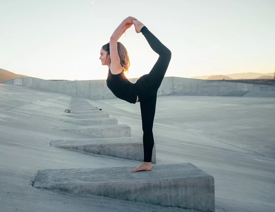 Eine Frau in schwarzer Sportkleidung nimmt auf einer Betonstruktur im Freien bei Sonnenuntergang eine stehende Yogapose ein und strahlt dabei die Zuversicht und Ruhe aus, die GLAMESiA auszeichnet.