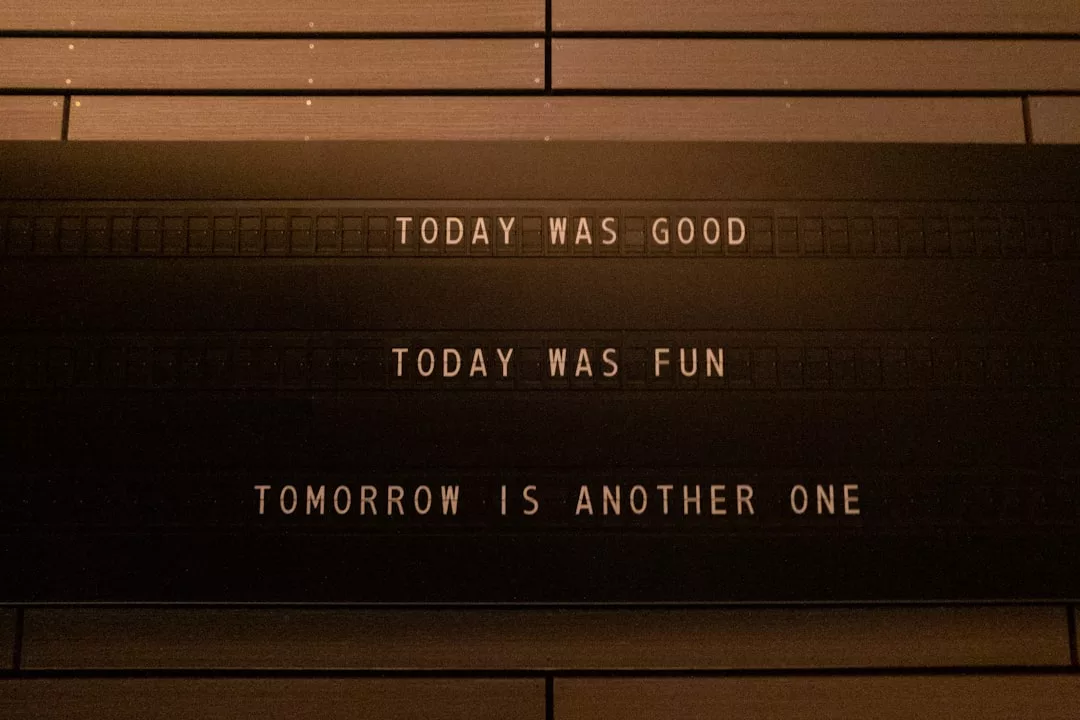 Auf einem schwarzen Schild mit weißen Buchstaben steht: "TODAY WAS GOOD, TODAY WAS FUN, TOMORROW IS ANOTHER ONE", das an einer Holzwand angebracht ist und dem Raum einen Hauch von GLAMESiA-Charme verleiht.