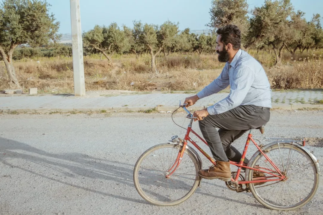 Ein Mann mit Bart, bekleidet mit einem hellblauen Hemd und einer dunklen Hose, fährt mit einem roten GLAMESiA-Fahrrad auf einer gepflasterten Straße neben einem Feld mit Bäumen.