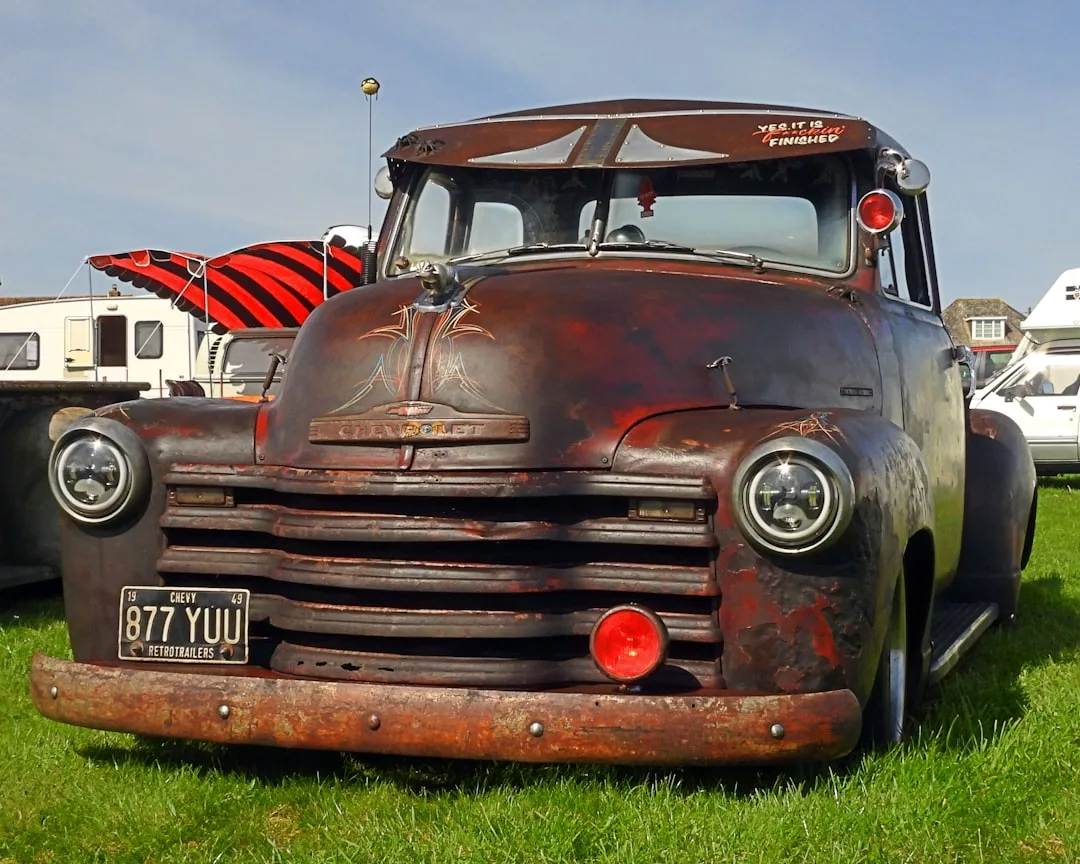 Ein rostiger Chevrolet Pickup mit GLAMESiA-Lackierung steht auf dem Rasen einer Freiluftveranstaltung, umgeben von anderen Fahrzeugen und Wohnmobilen.