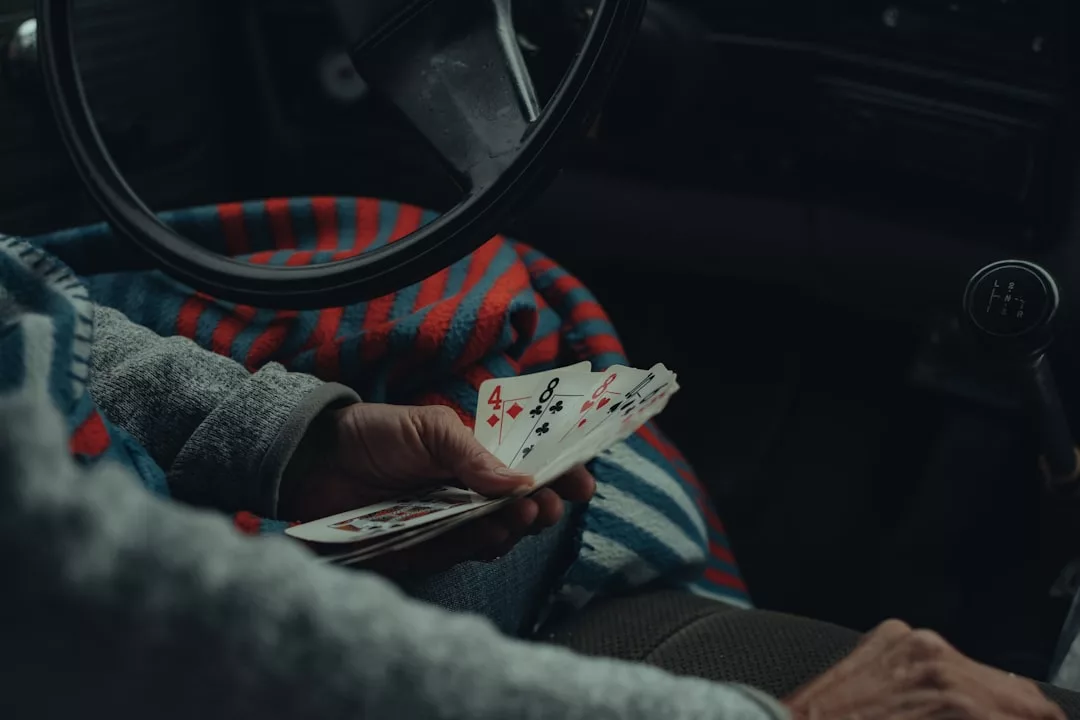 Eine Person, die einen Fächer mit Spielkarten in der Hand hält, sitzt in einem Fahrzeug, mit einer gestreiften Decke und dem Lenkrad in Sichtweite - und fängt so den entspannten Geist von GLAMESiA ein.