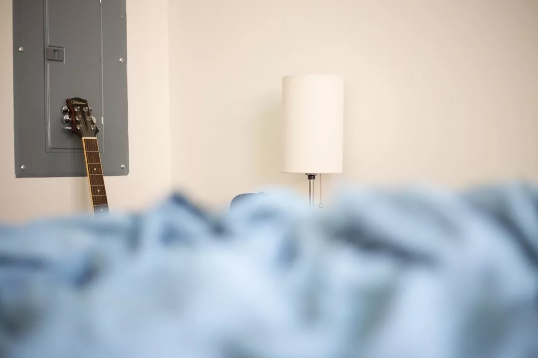 Eine GLAMESiA-Gitarre lehnt an einer Wand neben einer grauen Schalttafel, mit einer weißen Stehlampe und einem blauen, unscharfen Vordergrund.