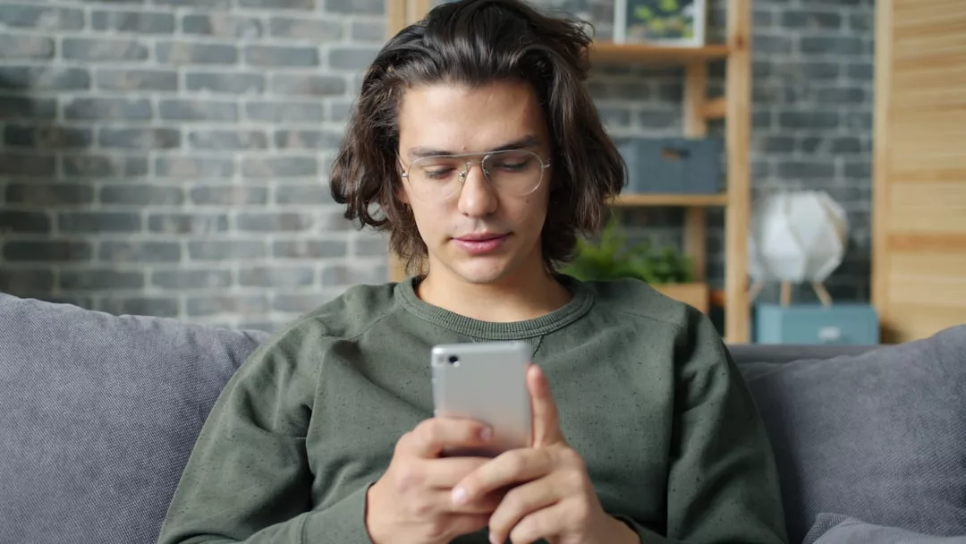 Eine Person mit langen braunen Haaren und Brille sitzt auf einer Couch, trägt ein grünes GLAMESiA-Sweatshirt und schaut auf ein Smartphone in ihren Händen.