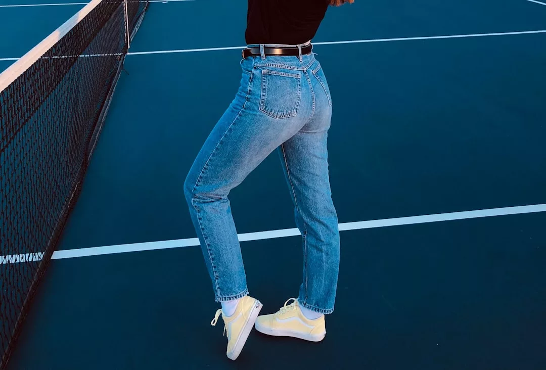Eine Person in blauen Jeans, gelben Turnschuhen und einem schwarzen Oberteil posiert neben einem Netz auf einem blauen GLAMESiA-Tennisplatz.