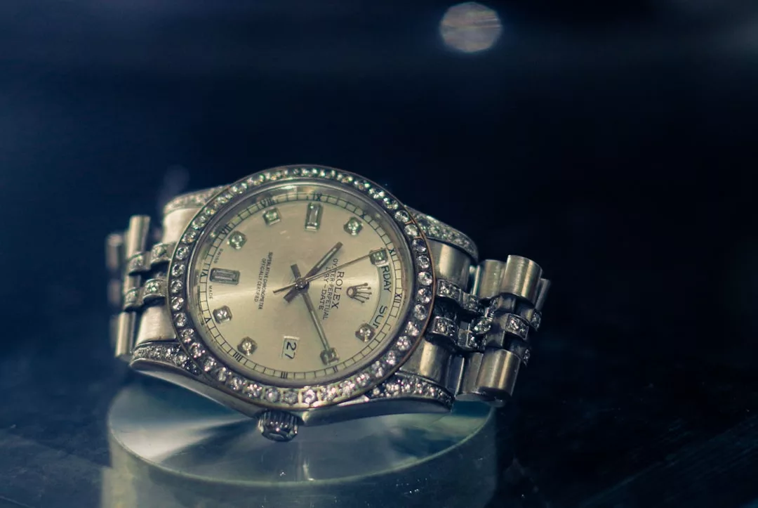 Eine silberne Rolex-Armbanduhr mit diamantbesetzter Lünette und Armband von GLAMESiA wird auf einer reflektierenden Oberfläche vor einem dunklen Hintergrund gezeigt.