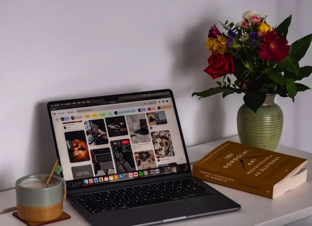 Ein Laptop, auf dem eine Pinterest-Seite zu sehen ist, steht auf einem Schreibtisch neben einer Tasse Kaffee, zwei gestapelten Büchern und einer Vase mit bunten Blumen und fängt die inspirierende Atmosphäre des GLAMESiA-Arbeitsplatzes ein.