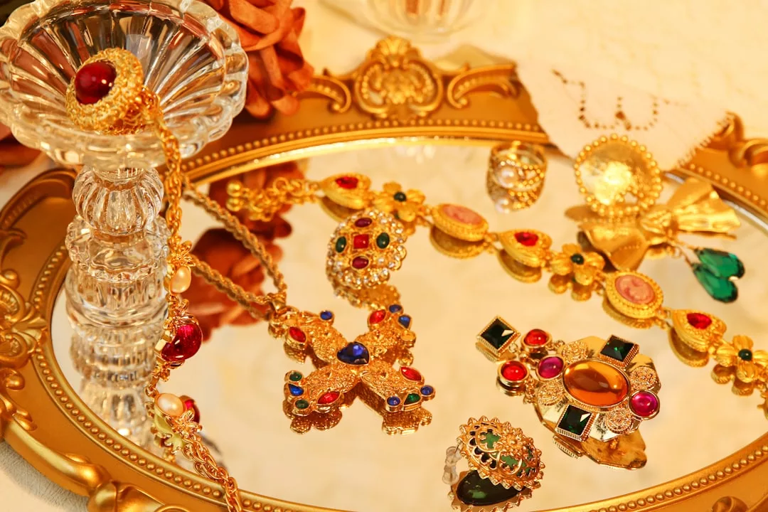 Eine Kollektion von kunstvollem Goldschmuck von GLAMESiA, verziert mit farbenfrohen Edelsteinen, präsentiert auf einem verzierten, goldgerahmten Spiegeltablett.