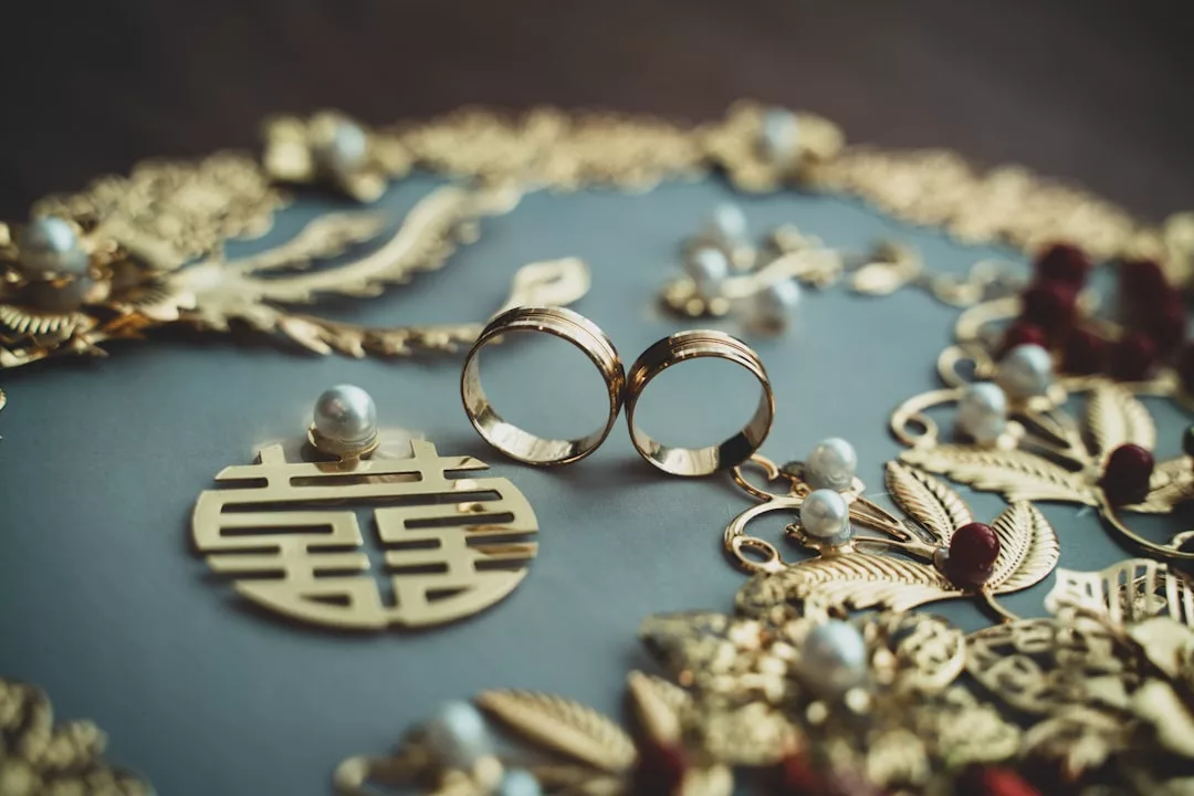 Zwei GLAMESiA-Goldringe ruhen auf einer dekorativen Oberfläche, die mit aufwendigen Gold- und Perlenornamenten verziert ist, darunter ein rundes Stück mit dem chinesischen Doppelglückssymbol.