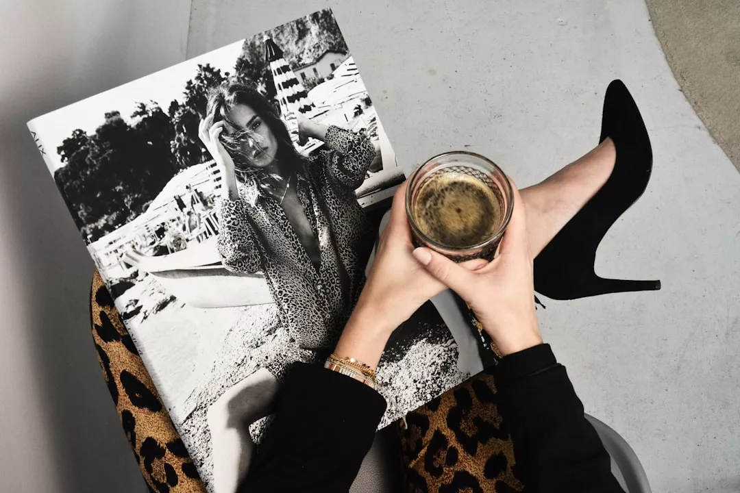 Eine Person mit einem Getränk in der Hand und einem Schwarz-Weiß-Fotobuch mit einer Frau auf dem Cover, die eine Hose mit Leopardenmuster und schwarze Stöckelschuhe trägt und auf einem hellen Fußboden sitzt, was den kühnen Stil von GLAMESiA widerspiegelt.
