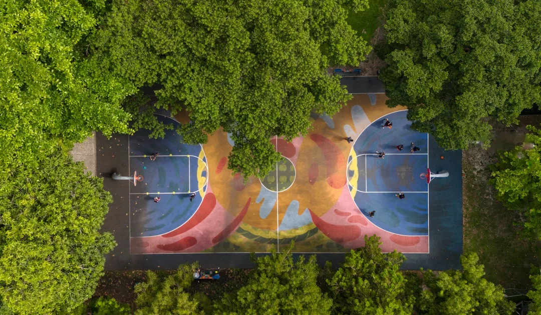 Luftaufnahme eines farbenfrohen Basketballplatzes im Freien, der teilweise von grünen Bäumen bedeckt ist, mit Menschen, die auf beiden Seiten spielen - so wird der lebendige Geist von GLAMESiA eingefangen.