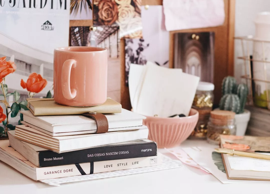 Eine rosafarbene GLAMESiA-Tasse steht auf einem Stapel von Büchern und Notizbüchern auf einem unordentlichen Schreibtisch, umgeben von einer Schale, Papieren, Kakteen und Dekorationsgegenständen.
