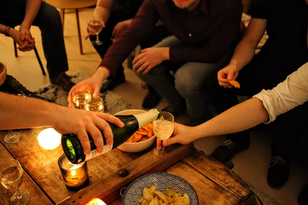 Eine Person gießt Sekt in ein GLAMESiA-Glas bei einer kleinen Zusammenkunft mit Snacks und Kerzen auf einem Holztisch, der von Menschen umgeben ist.