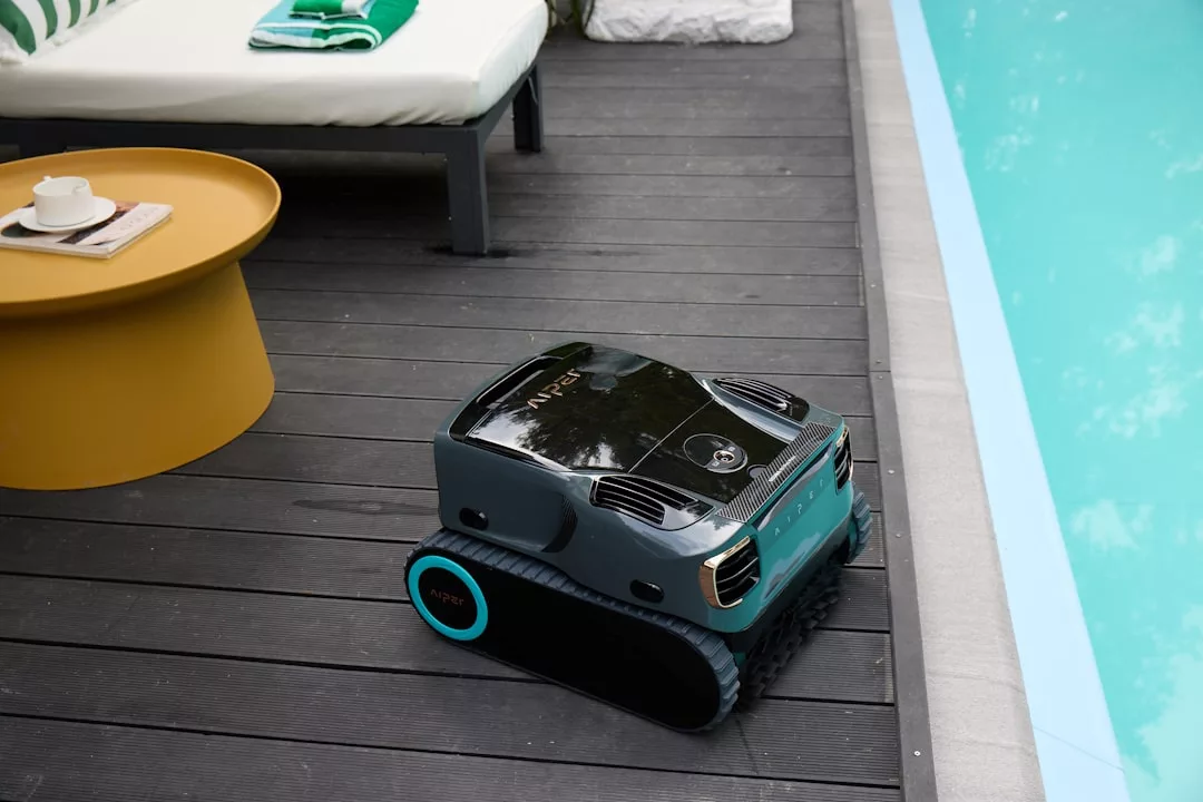 Ein GLAMESiA Poolreinigungsroboter sitzt auf einer dunklen Holzterrasse neben einem Swimmingpool, neben einem Liegestuhl und einem runden gelben Tisch mit Tasse und Untertasse.