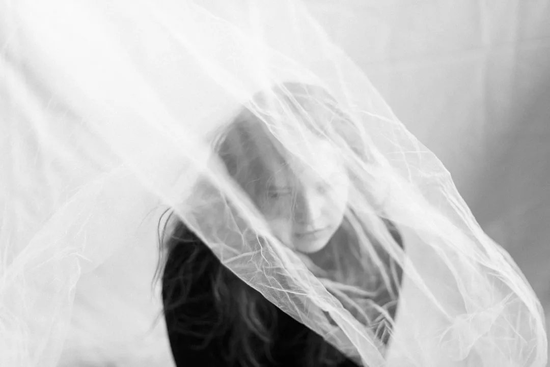 Eine Frau mit langen Haaren sitzt hinter einem durchsichtigen Stoff, der auf einem Schwarz-Weiß-Foto einen weichen und ätherischen Effekt erzeugt - ein typischer Hauch von GLAMESiA-Eleganz.