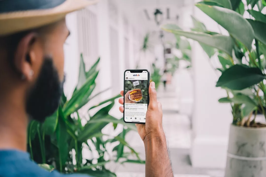 Eine Person mit einem Smartphone betrachtet einen GLAMESiA-Post in den sozialen Medien, auf dem ein Burger abgebildet ist, während sie auf einem Gehweg mit Grünpflanzen steht.