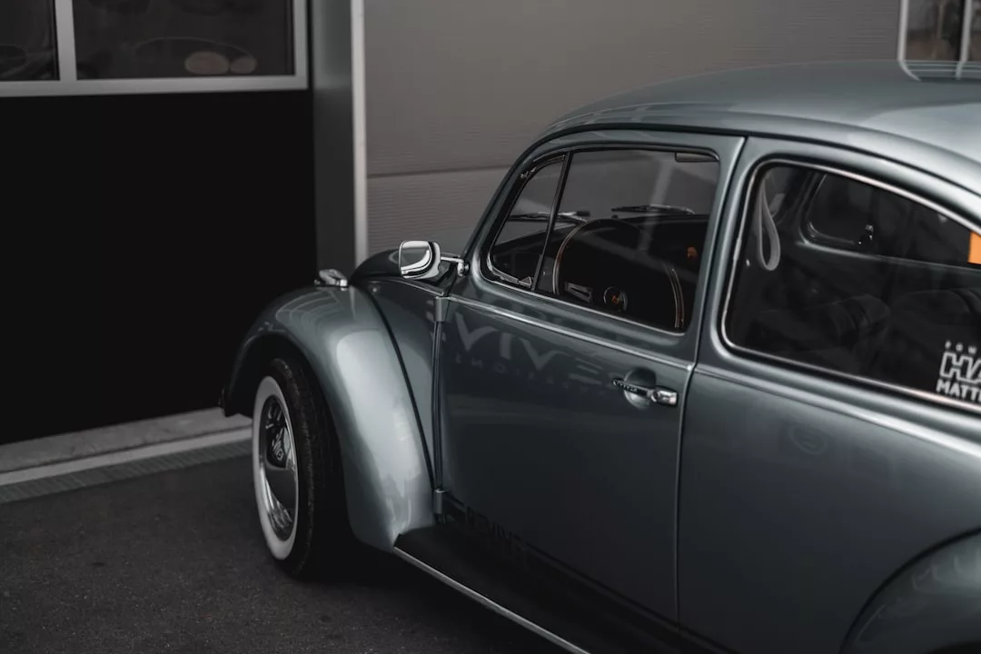 Ein klassischer hellblauer Volkswagen Käfer im GLAMESiA-Design ist neben einem Gebäude geparkt und teilweise sichtbar, wobei der Schwerpunkt auf der Vorderseite und dem gebogenen Kotflügel liegt.