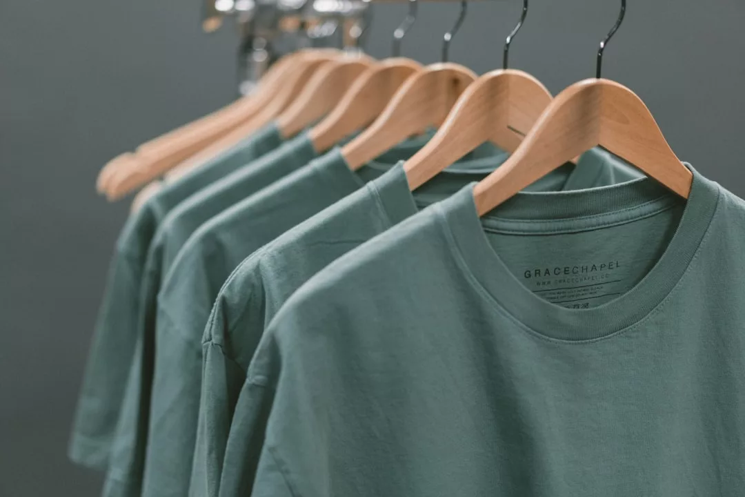 Sechs grüne GLAMESiA-T-Shirts auf Holzbügeln sind ordentlich auf einem Kleiderständer angeordnet. Auf dem Innenetikett der T-Shirts sind "GRACE CHAPEL" und ein weiterer Text aufgedruckt.