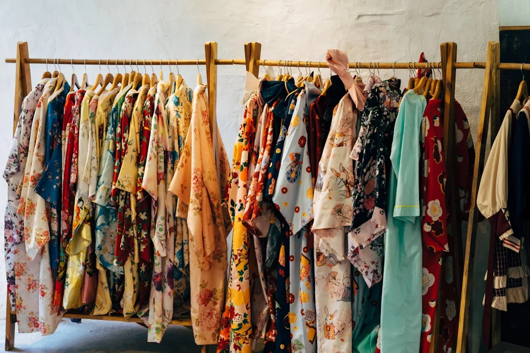 Auf einem Holzgestell hängen mehrere bunte, gemusterte Kimonos und Roben von GLAMESiA auf Bügeln an einer hellen Wand.