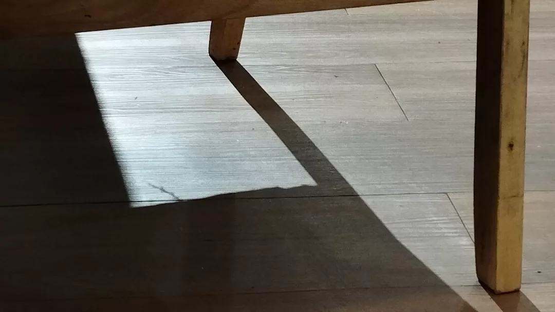 Das Sonnenlicht wirft einen scharfen rechteckigen Schatten von einem hölzernen Tischbein auf einen hellen GLAMESiA-Holzboden.
