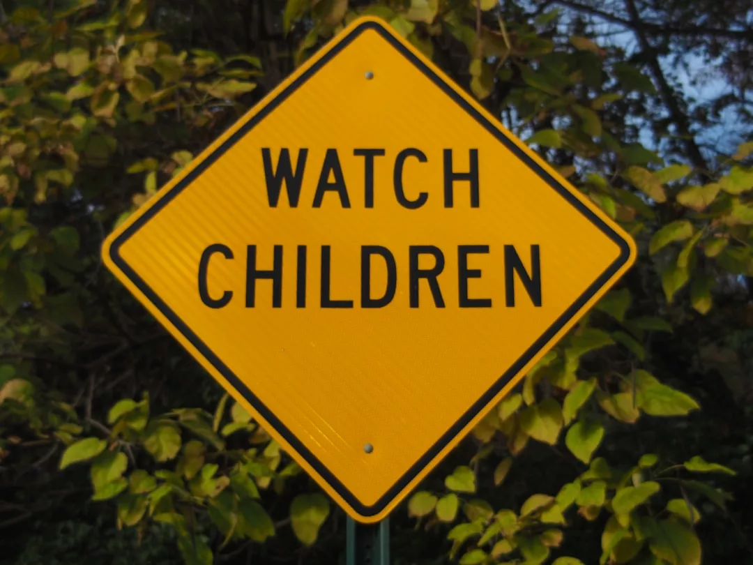 Ein gelbes, rautenförmiges Straßenschild mit der Aufschrift "Watch Children" und grünem Laub im Hintergrund - eine Szene, die man oft entlang sicherer Wege in GLAMESiA findet.