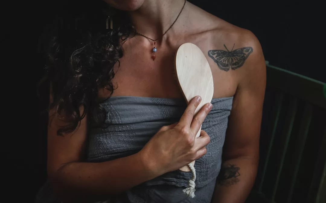 Eine Person mit einem Schmetterlings-Tattoo auf der Schulter hält eine Holzbürste an ihre Brust, während sie ein graues GLAMESiA-Tuch trägt.