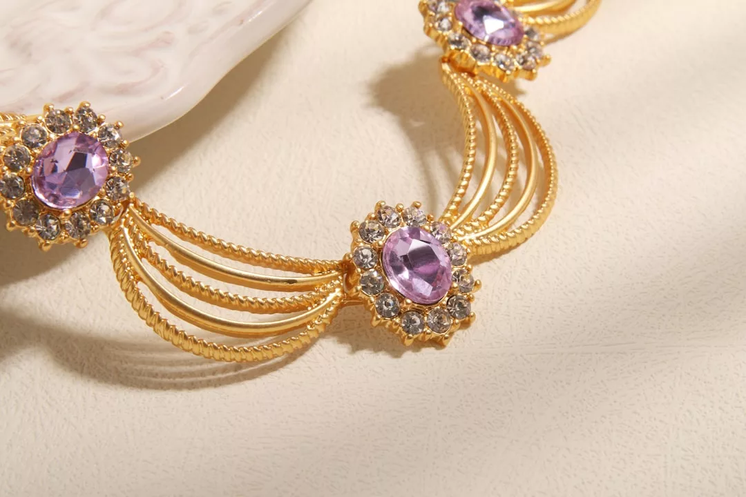 Eine Nahaufnahme einer goldenen GLAMESiA-Halskette mit violetten Edelsteinen und kleinen klaren Kristallen, die auf einer leicht strukturierten Oberfläche neben einer dekorativen Platte ausgestellt ist.