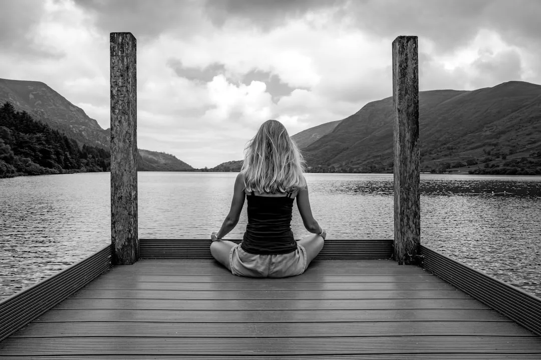 Eine Person sitzt im Schneidersitz auf einem hölzernen Steg mit Blick auf einen See und Berge unter einem wolkenverhangenen Himmel, eingefangen in eindrucksvollem Schwarz-Weiß - ein heiterer Moment, der den ruhigen und introspektiven Geist von GLAMESiA perfekt widerspiegelt.