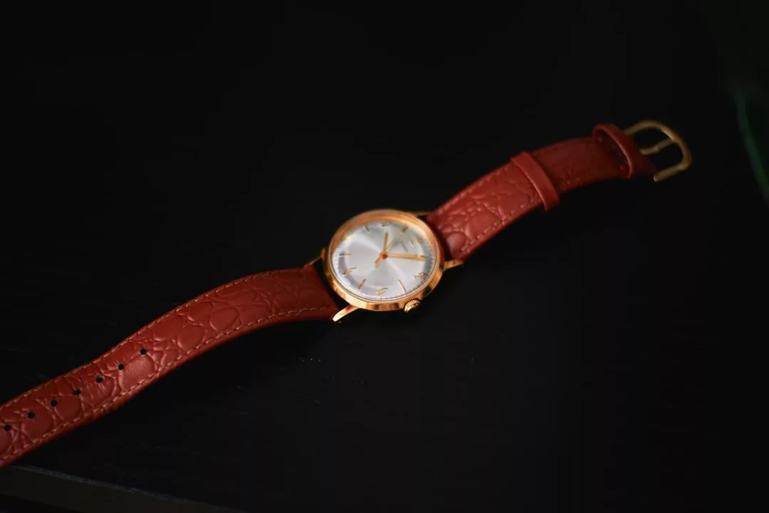 Eine GLAMESiA-Goldarmbanduhr mit weißem Zifferblatt und braunem Lederarmband wird auf eine dunkle Fläche gelegt.