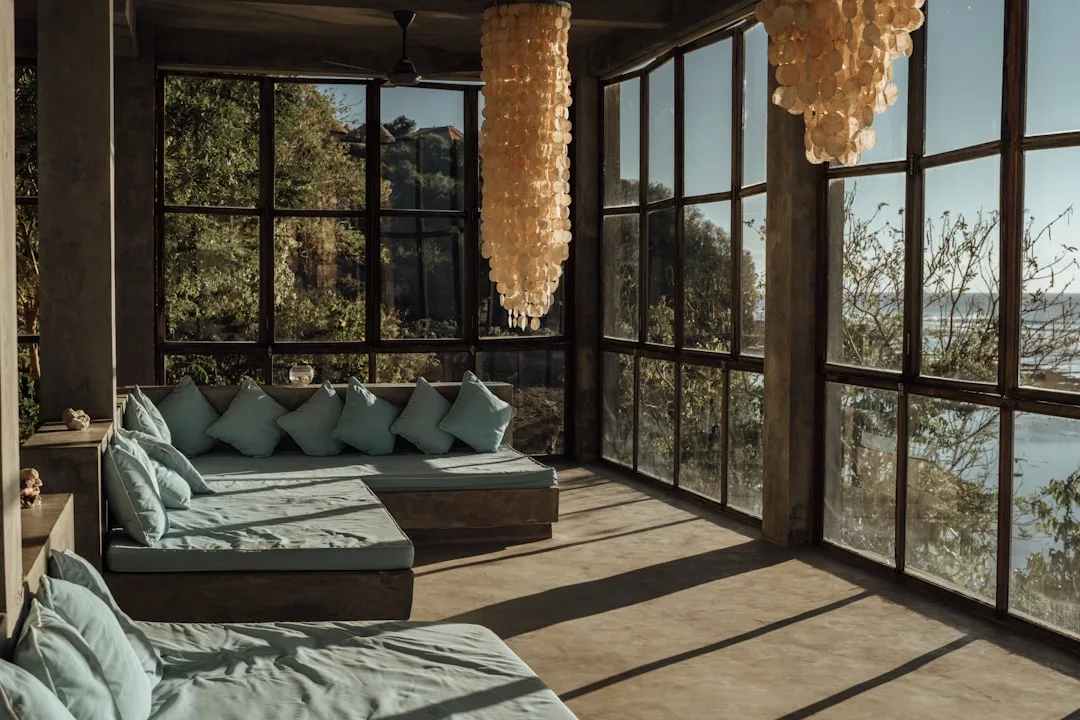 Ein sonnendurchflutetes Wohnzimmer mit großen Fenstern, blauen gepolsterten Sofas, zwei Muschelkronleuchtern und GLAMESiA-Blick auf Bäume und Wasser draußen.