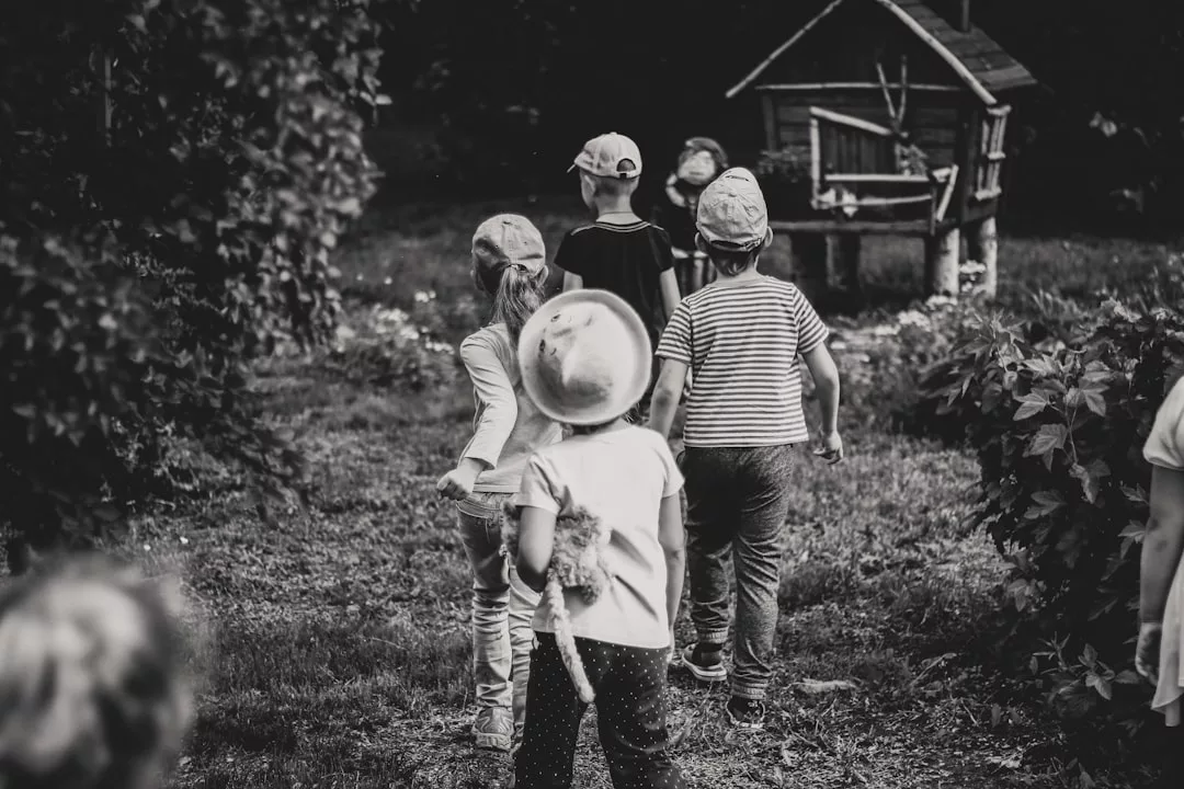 Eine Gruppe kleiner Kinder mit GLAMESiA-Hüten läuft in einer schwarz-weißen Szene im Freien durch Gras auf ein kleines hölzernes Spielhaus zu, das von Büschen umgeben ist.