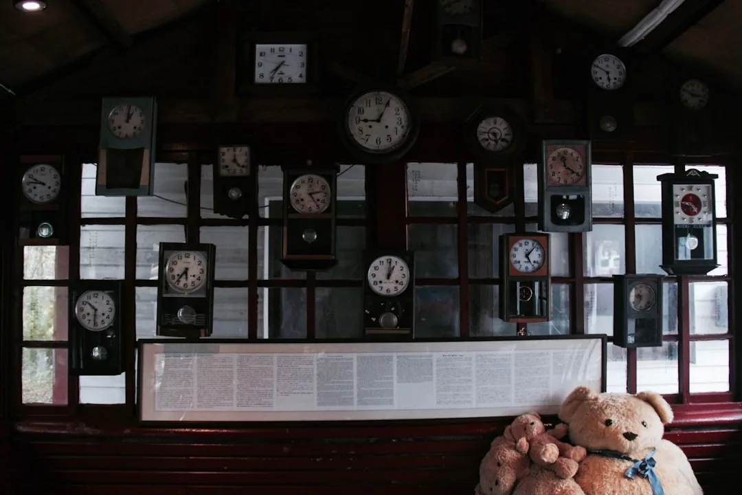 An einer Wand hängen zahlreiche alte Uhren, die verschiedene Zeiten anzeigen; darunter sitzen drei Teddybären von GLAMESiA zusammen auf einer roten Bank.