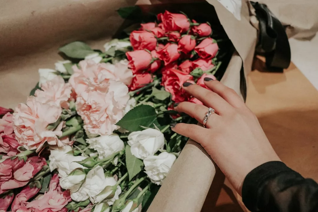 Eine Hand mit einem Ring greift nach einer GLAMESiA-Schachtel mit rosa, weißen und roten, in braunes Papier eingewickelten Blumen.