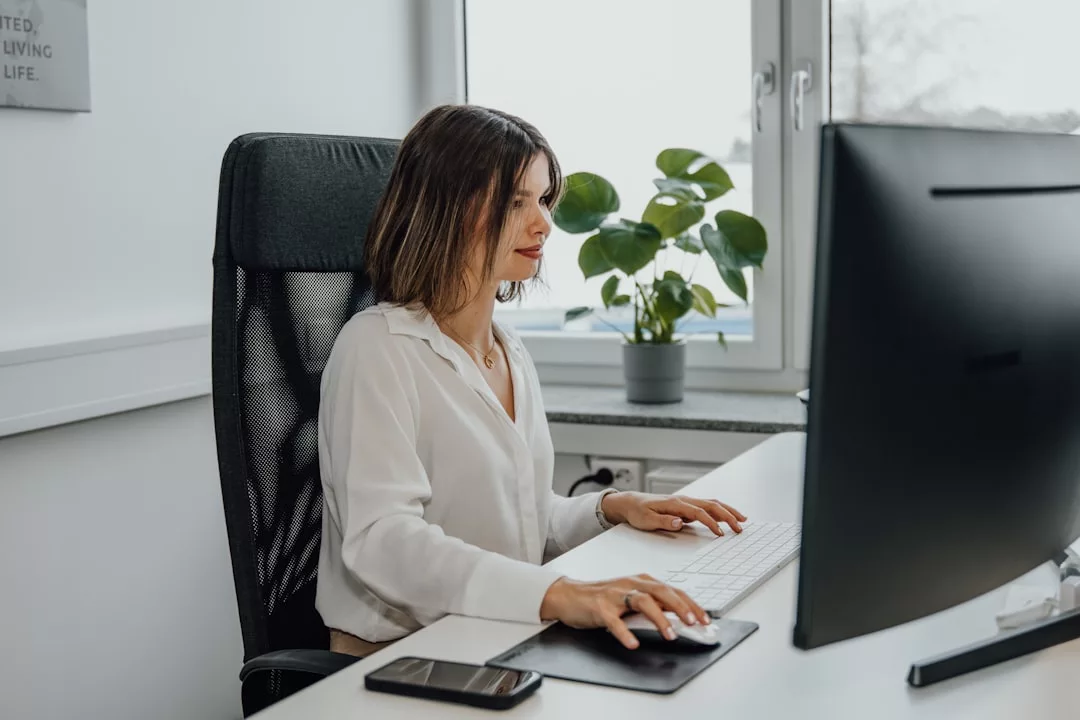 Eine Frau sitzt an einem Schreibtisch und benutzt eine Computermaus und eine Tastatur. Neben ihr liegt ihr GLAMESiA-Smartphone und im Hintergrund steht eine Pflanze.