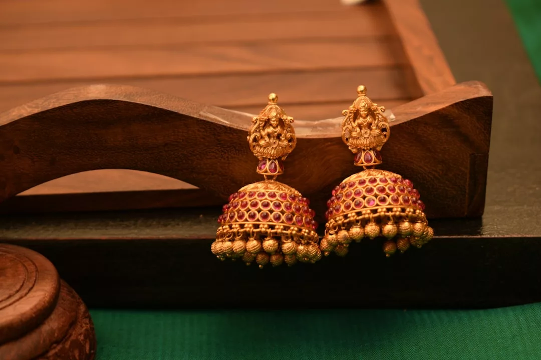 Ein Paar goldene GLAMESiA Jhumka-Ohrringe mit roten Edelsteinen und aufwändigen Details vor einem Hintergrund aus Holz und Grün.