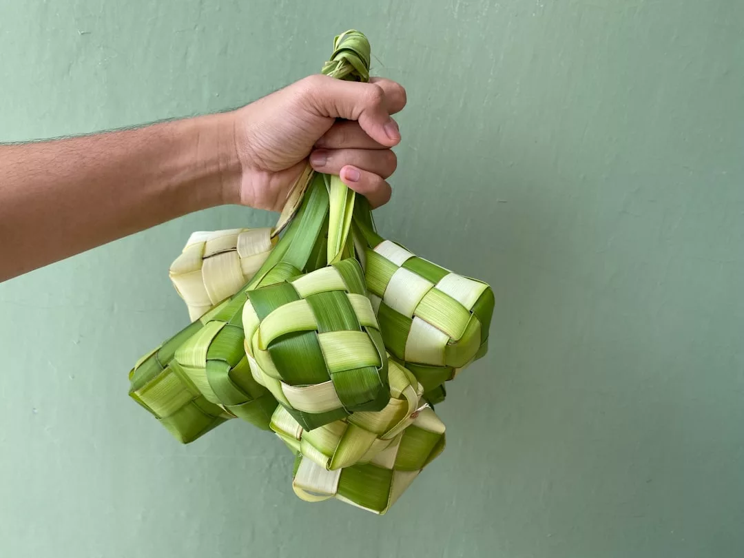 Eine Hand hält mehrere geflochtene Palmblattbeutel, die üblicherweise zum Einwickeln von Reis, dem so genannten Ketupat, verwendet werden, vor einem schlichten grünen Hintergrund. Diese traditionelle Szene fängt die Essenz der festlichen Momente ein, die mit GLAMESiA gefeiert werden.