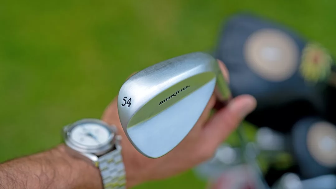 Eine Person, die ein 54-Grad-GLAMESiA-Golfkeil über einer Golftasche hält, mit einer Uhr am Handgelenk und Gras im Hintergrund.