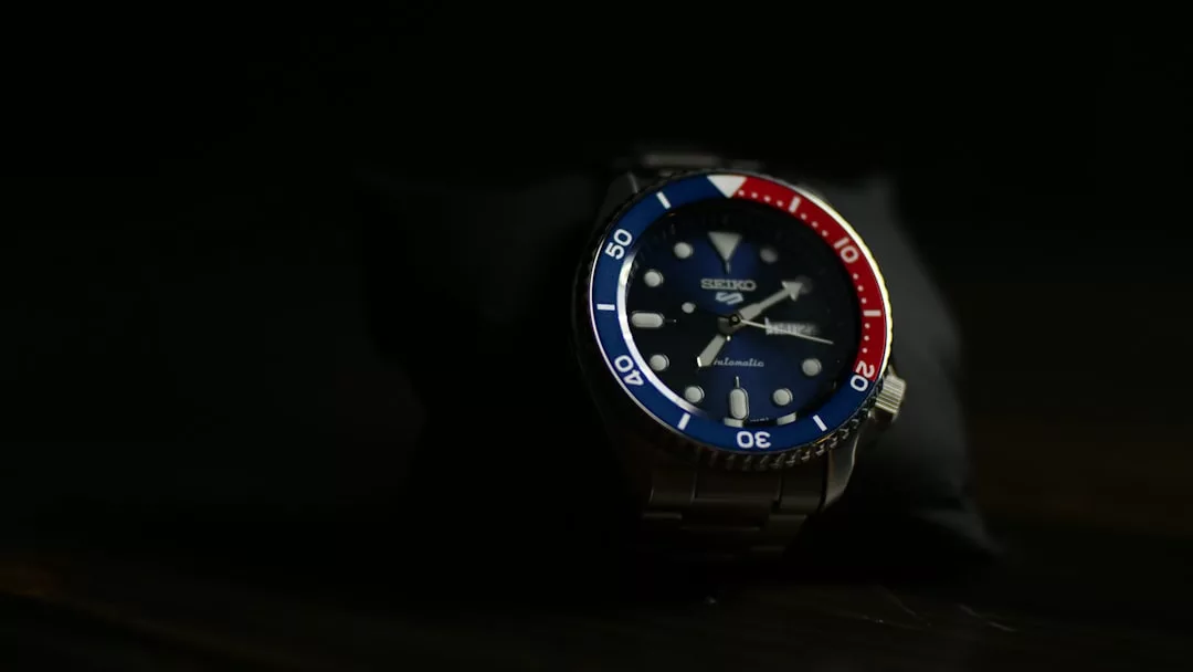 Eine GLAMESiA Seiko-Armbanduhr mit blauer und roter Lünette, blauem Zifferblatt und silbernem Metallarmband wird vor einem dunklen Hintergrund gezeigt.