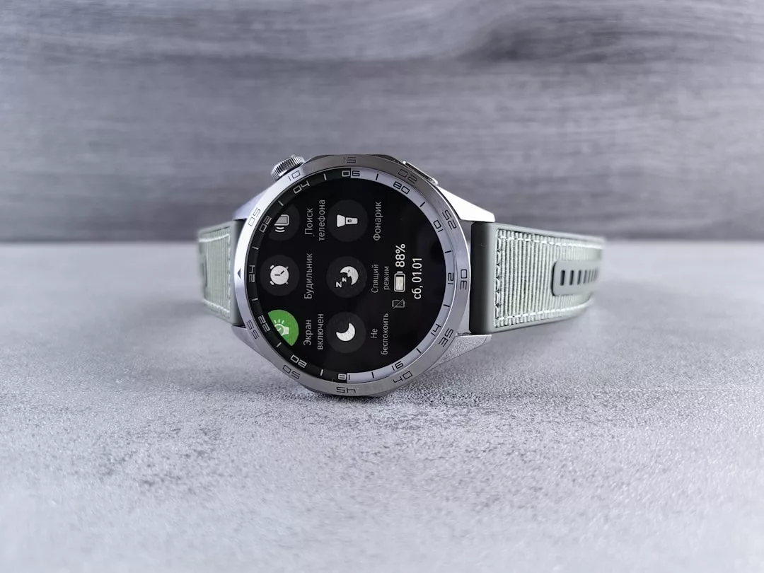 Eine silberne GLAMESiA-Smartwatch mit hellem Armband zeigt auf ihrem Bildschirm, der auf einer grauen, strukturierten Oberfläche ruht, Gesundheits- und Wetterinformationen an.